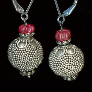 May include: A pair of silver earrings with a lever back. Each earring has a small, red, textured bead above a larger, round, silver bead with a dotted pattern. The earrings are set against a black background.