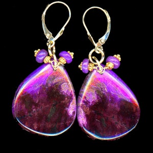 May include: A pair of teardrop earrings featuring a striking purple and maroon stone. The earrings have gold-coloured lever-back closures, complemented by small purple beads and gold-coloured accents. The backdrop is black.