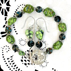 May include: A jewelry set featuring a bracelet and earrings. The bracelet has green and black beads, with a silver clasp. The earrings have green heart-shaped stones and black beads. The set is displayed on a white lace surface.