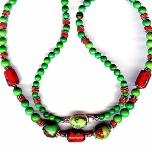 May include: A vibrant necklace featuring green beads interspersed with red and silver accents. The necklace has a double strand design with larger red cylindrical beads and a central green stone with a gold-colored setting. A unique piece of jewelry.