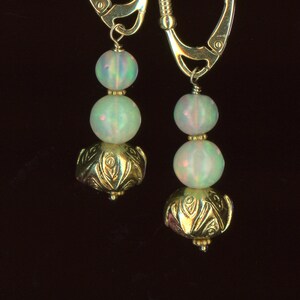 May include: A pair of gold-colored leverback earrings. Each earring features two opalescent, round beads and a decorative gold-colored bead. The earrings hang from a gold-colored leverback clasp.