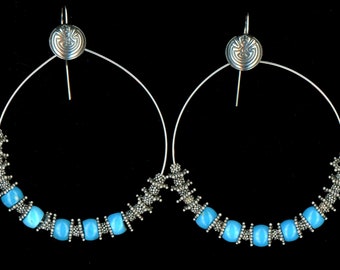 May include: Silver hoop earrings with a spiral design and light blue beads. The earrings have a circular design with a silver hook and a decorative silver element at the top. The lower portion of the hoops are adorned with light blue beads.