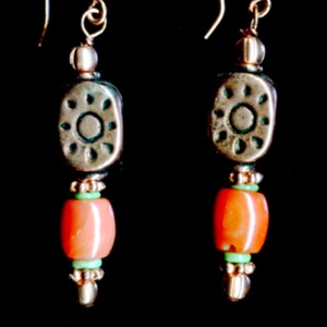 May include: A pair of handcrafted earrings with a dark background. Each earring features a gold-colored hook, a silver-toned bead, and a decorative oval-shaped bead with a floral design. Below, there is a coral-colored bead, a small green bead, and a cluster of silver beads.
