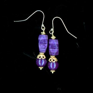 May include: A pair of purple earrings with gold-colored accents. Each earring features a rectangular purple stone, a gold-colored decorative element, and a round purple bead. The earrings hang from silver-colored ear wires.