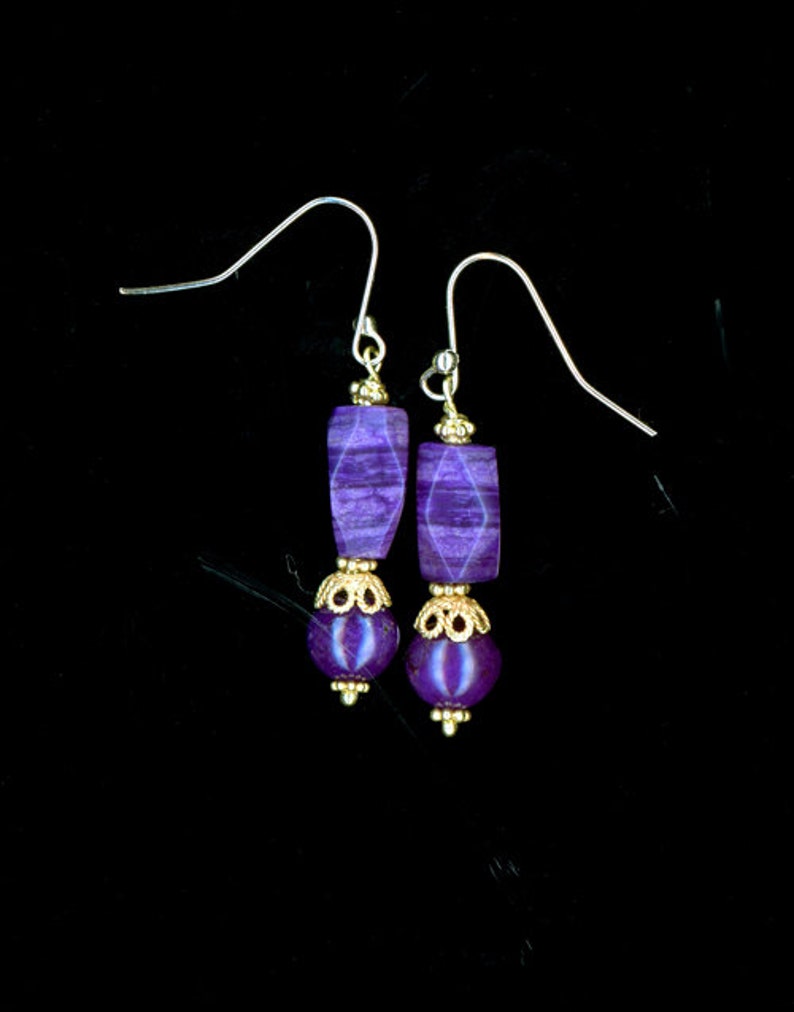 May include: A pair of purple earrings with gold-colored accents. Each earring features a rectangular purple stone, a gold-colored decorative element, and a round purple bead. The earrings hang from silver-colored ear wires.
