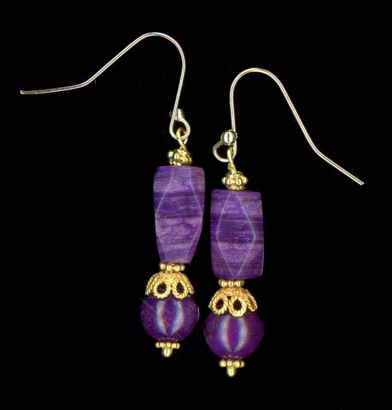 May include: A pair of dangle earrings with purple gemstone beads and gold-colored accents. The earrings feature faceted rectangular and round beads, connected by ornate gold-colored metalwork and silver-colored ear wires. The background is black.