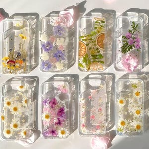 May include: A collection of clear phone cases, each embedded with dried flowers and botanical elements. The cases feature various floral arrangements, including daisies, lavender, and orange slices, creating a unique and artistic aesthetic. The cases are arranged on a white surface with scattered pink petals.