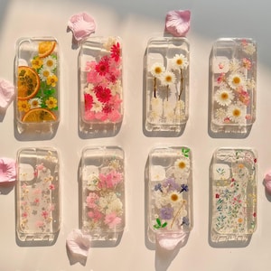 Puede incluir: Ocho fundas transparentes para teléfono con diferentes diseños de flores prensadas. Las fundas están dispuestas en dos filas de cuatro. Los diseños incluyen una variedad de flores, como margaritas, rosas y flores silvestres. Las fundas son todas transparentes y muestran las flores en detalle.