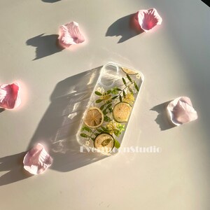 May include: Clear phone case with dried lemon slices, small yellow flowers, and green leaves. The case is rectangular and has a transparent design. Pink flower petals are scattered around the case on a white surface. The text "EvermoonStudio" is visible.