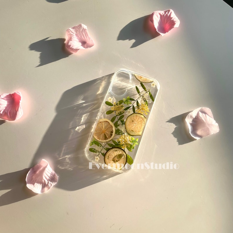 May include: Clear phone case with dried lemon slices, small yellow flowers, and green leaves. The case is rectangular and has a transparent design. Pink flower petals are scattered around the case on a white surface. The text "EvermoonStudio" is visible.