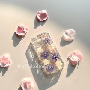 May include: Clear phone case with embedded purple, white, and pink dried flowers. The case is surrounded by scattered pink flower petals. The text "SkyLakeDesign" is visible on the surface.