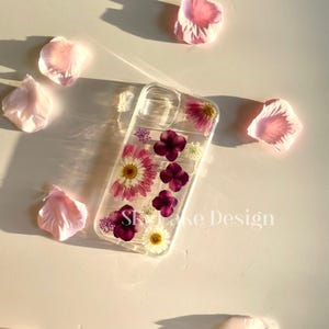 May include: Clear phone case with embedded dried flowers, including pink daisies and purple blossoms. The case has the text "Skylake Design" and is surrounded by scattered pink flower petals.