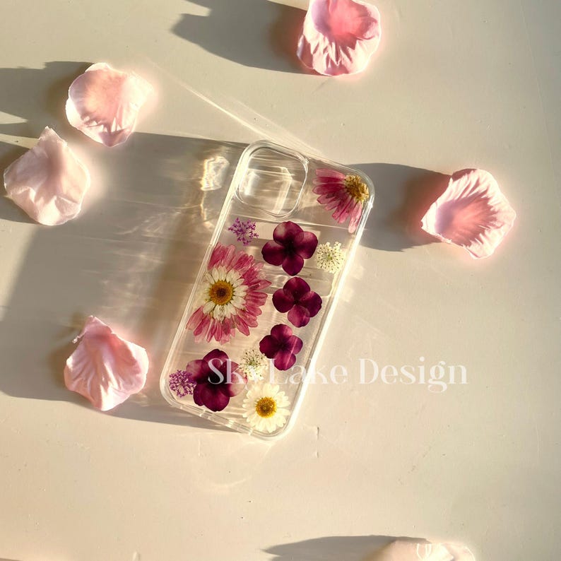 May include: Clear phone case with embedded dried flowers, including pink daisies and purple blossoms. The case has the text "Skylake Design" and is surrounded by scattered pink flower petals.