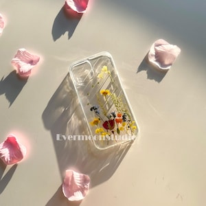 May include: Clear phone case with dried flowers in yellow, red, and blue. The case is rectangular with rounded corners and has the text "Evermoonstudio" printed on it. Pink flower petals are scattered around the case.