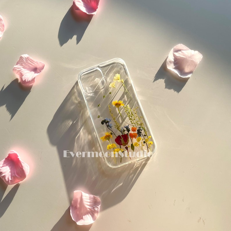 May include: Clear phone case with dried flowers in yellow, red, and blue. The case is rectangular with rounded corners and has the text "Evermoonstudio" printed on it. Pink flower petals are scattered around the case.