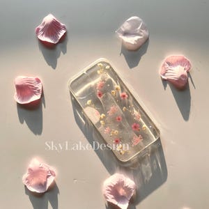 May include: A clear rectangular phone case with embedded pink and yellow dried flowers. The case is surrounded by scattered pink flower petals. The text "SkyLakeDesign" is visible on the surface.
