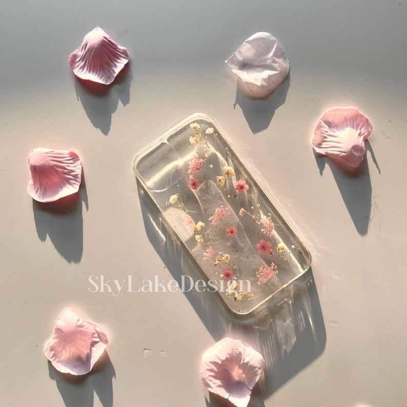 May include: A clear rectangular phone case with embedded pink and yellow dried flowers. The case is surrounded by scattered pink flower petals. The text "SkyLakeDesign" is visible on the surface.