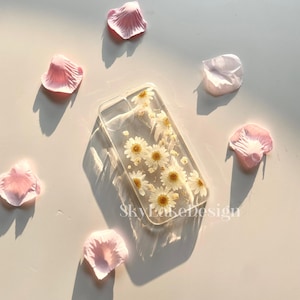 May include: Clear phone case with embedded white daisies and yellow centers. The case is rectangular and features the text "SkyLakeDesign". Pink flower petals are scattered around the case on a light surface.