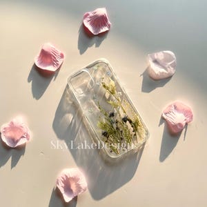 May include: Clear phone case with dried flowers and the text "SkyLakeDesign". The case features a floral arrangement with blue and green accents. Pink petals are scattered around the case.