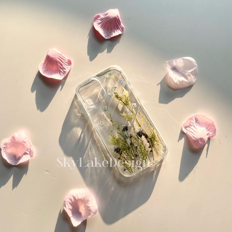 May include: Clear phone case with dried flowers and the text "SkyLakeDesign". The case features a floral arrangement with blue and green accents. Pink petals are scattered around the case.
