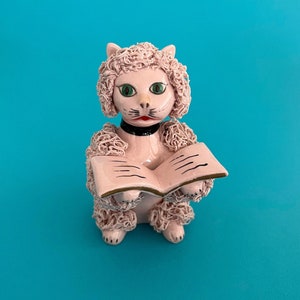 Może przedstawiać: Ceramiczna figurka kota z jasnoróżowym szkliwionym wykończeniem i teksturowanymi, kręconymi detalami futra. Kot ma jasne zielone oczy, czarną obrożę i trzyma otwartą różową książkę z czarnymi liniami na stronach.