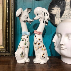 Może przedstawiać: Dwie białe ceramiczne figurki psów, jedna pudla i jedna teriera, z kwiatowymi wzorami i niebieskimi i różowymi kokardkami. Figurki stoją na drewnianej powierzchni.