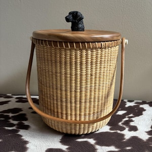 May include: Woven wicker basket with a wooden lid and handle. The lid is topped with a black dog figurine. The basket has a light brown color and a rounded shape. The handle is made of wood and is attached to the sides of the basket.