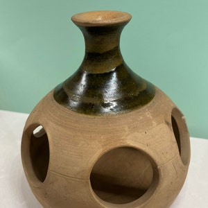 May include: A terracotta pot with a brown glaze and circular cutouts. The pot has a narrow neck and a rounded body.