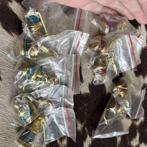 Może przedstawiać: Kolekcja małych, złotych brelok&oacute;w w przezroczystych plastikowych woreczkach. Niekt&oacute;re breloki mają zielone akcenty, inne czerwone. Breloki leżą na brązowo-beżowej powierzchni.
