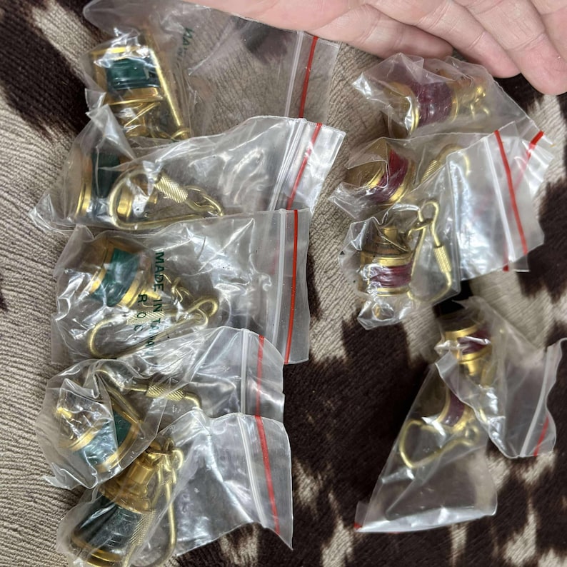 Może przedstawiać: Kolekcja małych, złotych brelok&oacute;w w przezroczystych plastikowych woreczkach. Niekt&oacute;re breloki mają zielone akcenty, inne czerwone. Breloki leżą na brązowo-beżowej powierzchni.