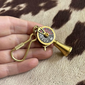 May include: A gold-colored keychain featuring a miniature ship's wheel and a brass-colored horn. The wheel has black and red details with directional labels. The keychain includes a spring-loaded clasp for easy attachment.