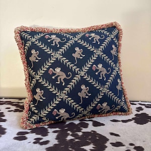 May include: Square decorative pillow with a navy blue background featuring a repeating pattern of monkeys holding fruit and leafy vines. The pillow has a red fringe border.