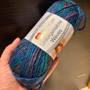 May include: A skein of Red Heart Boutique Unforgettable Waves yarn in a variegated blend of deep blue, teal, and dark purple tones. The white paper label displays the product name, 100% acrylic content, and a net weight of 3.5 ounces.