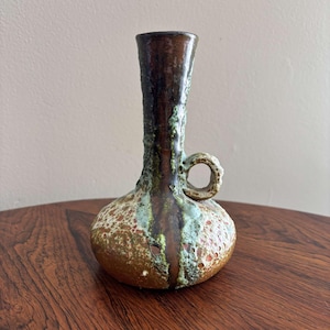 May include: A small, brown and green ceramic vase with a handle. The vase has a speckled glaze and a textured surface.