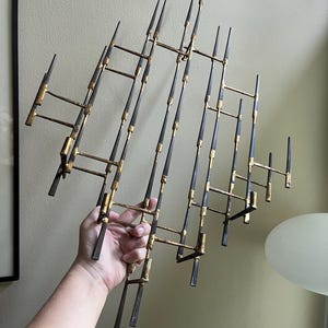 May include: A large, abstract wall sculpture made of metal. The sculpture is composed of many thin, pointed rods that are connected together in a geometric pattern. The rods are painted in a combination of black and gold.