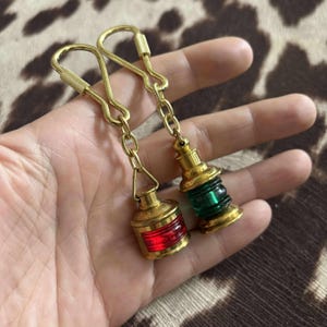 May include: Two brass keychains featuring miniature lantern charms. One lantern has a red lens, the other a green lens. Both are attached to a gold-colored chain and a clasp. The keychains are held in a hand.