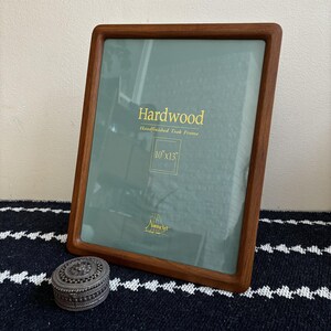 May include: A rectangular hardwood picture frame with a hand-finished teak frame. The frame holds a 10" x 13" image. The text "Hardwood Handfinished Teak Frame" is printed on the glass. A small, decorative silver box sits in front of the frame.