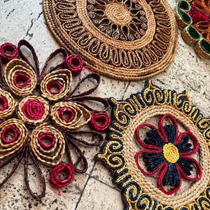 May include: Three circular woven mats featuring intricate coiled designs. One mat displays tan and dark brown patterns, another features tan and magenta loops, and the third showcases a central yellow flower with black petals and a tan border.