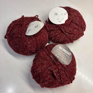 Può includere: Tre gomitoli di lana rosso scuro con una sottile trama mélange. Ogni gomitolo presenta un'etichetta di carta circolare. Un'etichetta riporta chiaramente il testo BONGIO, 90% WOOL - 10% VISCOSE, 50 g - 112 m, 1.78 oz - 123 yds e icone di lavaggio.