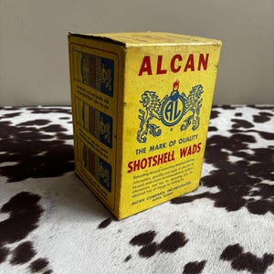 May include: A yellow ALCAN Shotshell Wads box with blue and red text and graphics. The box has text that reads "THE MARK OF QUALITY SHOTSHELL WADS" and "ALCAN COMPANY, INCORPORATED ALTON, ILLINOIS."
