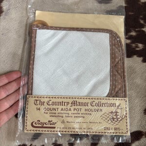 May include: A packaged pot holder kit from The Country Manor Collection. The kit includes a white Aida cloth, a brown patterned border, and a wooden handle. The packaging includes text that reads "14 Count Aida Pot Holder" and "For cross stitching, candle wicking, stencilling, fabric painting."