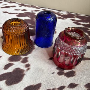 May include: Three glass light shades in different colors and textures. One is amber with a diamond pattern, one is cobalt blue with a smooth surface and embossed floral details, and one is a deep red with a diamond-cut top and smooth base.