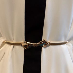 May include: A thin, metallic gold-tone belt featuring a central bow-shaped buckle. The belt is worn over a white garment with a prominent vertical black stripe running down the center, creating a high-contrast look with the polished gold accessory.