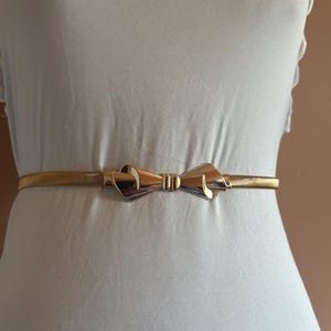 May include: A thin, gold-toned metallic belt featuring a central bow-shaped buckle. The belt has a flexible, coiled wire construction that wraps around the waist, with the polished gold bow serving as the primary decorative focal point.