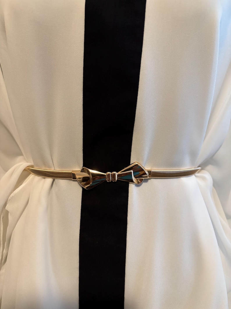 May include: A thin, metallic gold-tone belt featuring a central bow-shaped buckle. The belt is worn over a white garment with a prominent vertical black stripe running down the center, creating a high-contrast look with the polished gold accessory.
