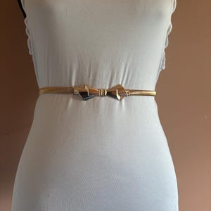 May include: A thin, metallic gold-toned belt featuring a central bow-shaped buckle. The belt is worn around the waist over a plain white garment. The buckle has a polished, reflective surface with a smooth, symmetrical design at the center.