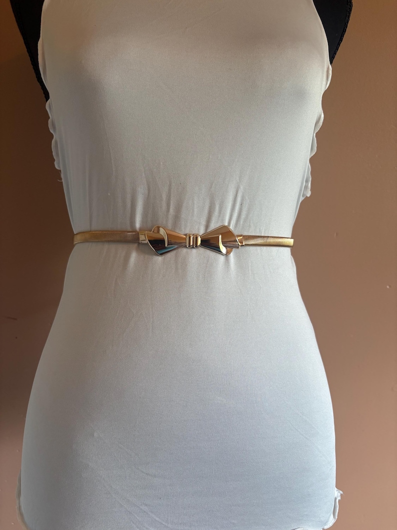 May include: A thin, metallic gold-toned belt featuring a central bow-shaped buckle. The belt is worn around the waist over a plain white garment. The buckle has a polished, reflective surface with a smooth, symmetrical design at the center.