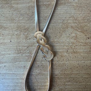 May include: A thin, flexible gold-tone metal belt featuring a textured, coiled band and a central decorative clasp composed of two interlocking, polished gold-tone metal links that create a smooth, reflective, and continuous loop design.
