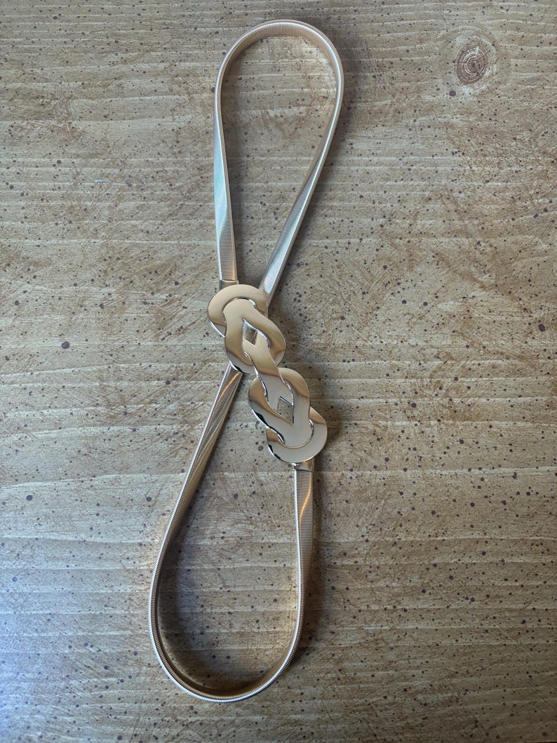 May include: A thin, flexible gold-tone metal belt featuring a textured, coiled band and a central decorative clasp composed of two interlocking, polished gold-tone metal links that create a smooth, reflective, and continuous loop design.