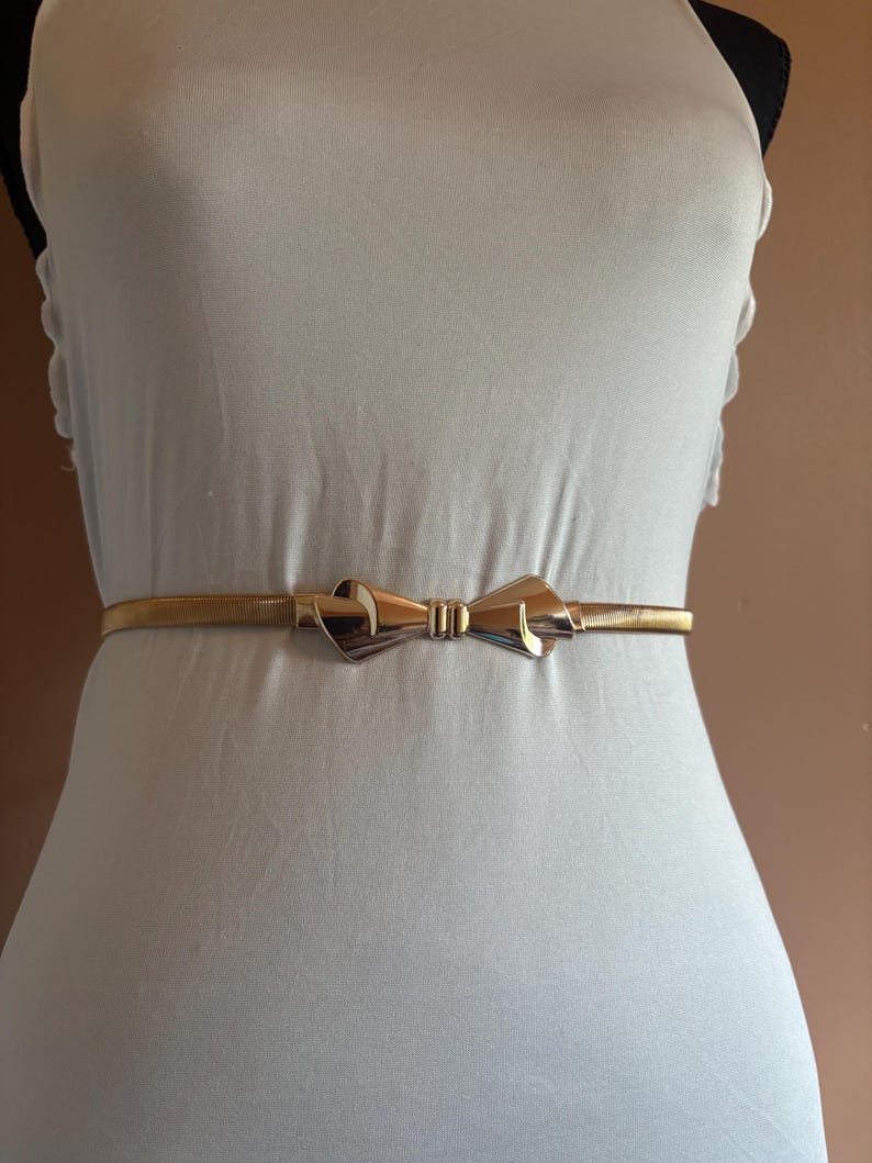 May include: A thin, gold-toned metallic belt featuring a central bow-shaped buckle. The belt has a flexible, coiled wire construction that wraps around the waist, with the polished gold bow serving as the primary decorative focal point.